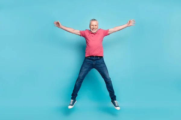 Фотография в полный рост, на которой бородатый мужчина в розовой футболке джинсах улыбается, прыгая высоко на голубом фоне — стоковое фото