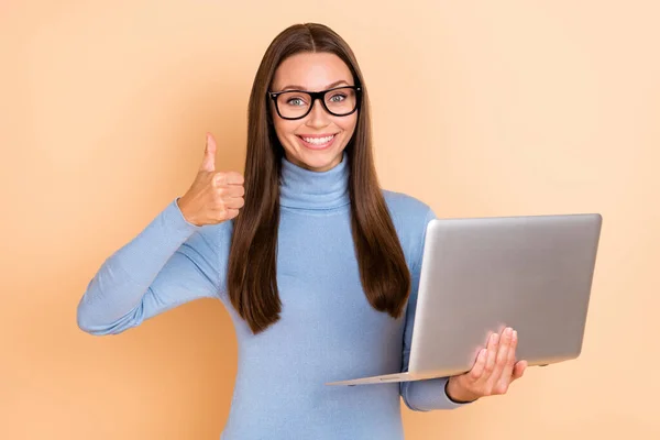 Photo de jolie brune jeune femme tenir ordinateur portable pouce vers le haut porter lunettes col roulé bleu isolé sur fond de couleur beige — Photo