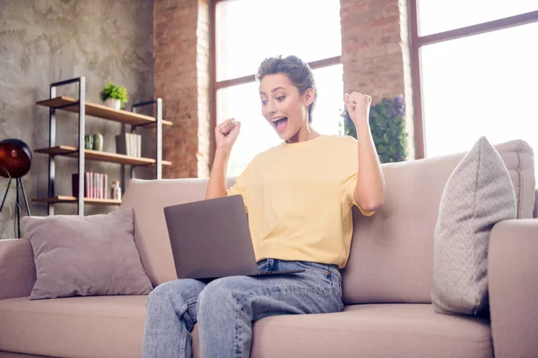 Photo of positive joyful lady use pc świętować zwycięstwo siedzieć sofa otwarte usta nosić żółty t-shirt w nowoczesnym domu w pomieszczeniach — Zdjęcie stockowe