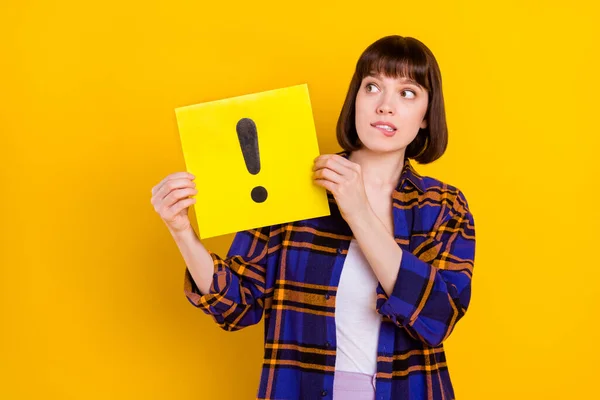 Retrato de menina intrigante atraente segurando placa de sinal de exclamação mordendo lábio isolado sobre fundo de cor amarela brilhante — Fotografia de Stock