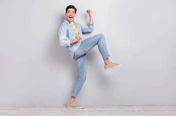 Foto van schattige gelukkige jonge man gekleed denim shirt lopen lege ruimte stijgende vuisten geïsoleerde beton grijze muur achtergrond — Stockfoto