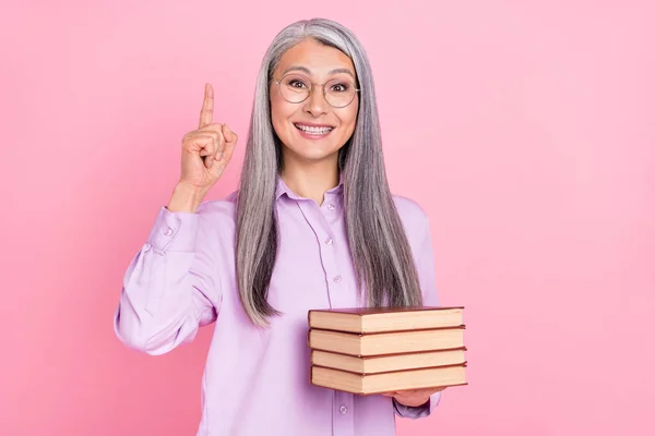 Retrato de mulher de cabelos grisalhos alegre atraente segurando livros apontando para cima encontrar solução isolada sobre fundo cor pastel rosa — Fotografia de Stock
