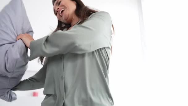 Позитивная женщина танцует на мягкой кровати бросать подушки носить тонкий пижаму в удобной комнате — стоковое видео