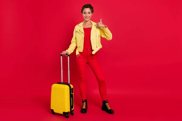 Фотография блестящей милой молодой женщины, одетой в кожаную куртку с багажом, показывающей большой палец вверх, улыбающийся красный цвет фона — стоковое фото