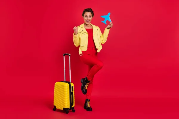 魅力的な幸運な若い女性の写真は、青い航空機の手荷物を保持笑みを浮かべて黄色のジャケットを身に着けている拳隔離された赤い色の背景 — ストック写真