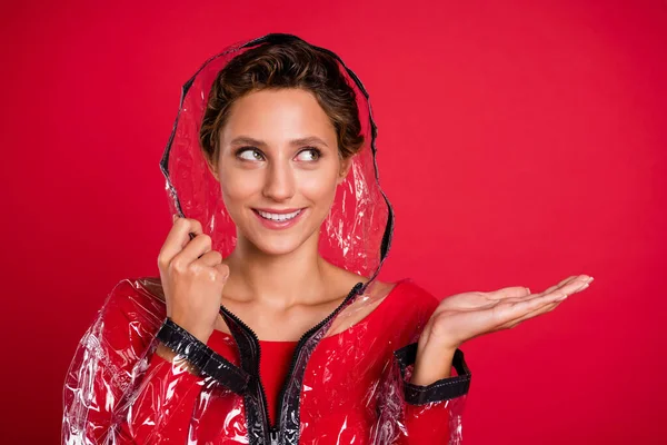 Foto de brilhante muito jovem mulher vestida casaco de borracha braço capuz sorrindo segurando braço olhando espaço vazio isolado cor vermelha fundo — Fotografia de Stock