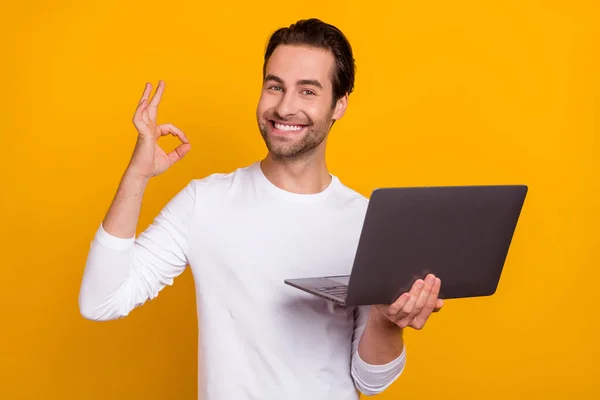 Zdjęcie fajnego brunet młody facet trzymać laptop pokazać okey nosić biały strój odizolowany na jasnożółtym tle kolor — Zdjęcie stockowe