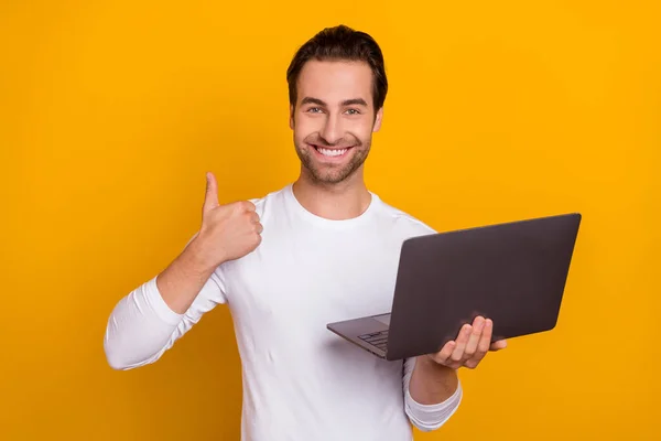 Zdjęcie optymistycznego brunet milenijny facet trzymać laptop pokazać kciuk do góry nosić biały strój izolowany na jasnożółtym tle kolor — Zdjęcie stockowe