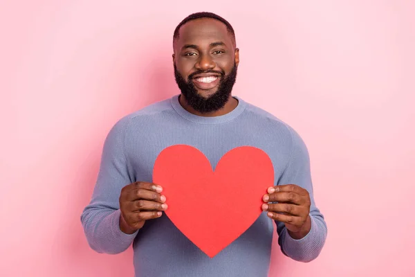 Foto de chico romántico alegre celebrar corazón rojo símbolo sonrisa dentada desgaste camisa gris aislado color rosa fondo — Foto de Stock