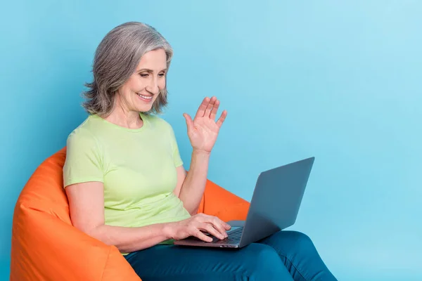 Photo portrait femme aînée travaillant sur un ordinateur portable assis dans un sac de fèves agitant main isolé fond de couleur bleu pastel — Photo