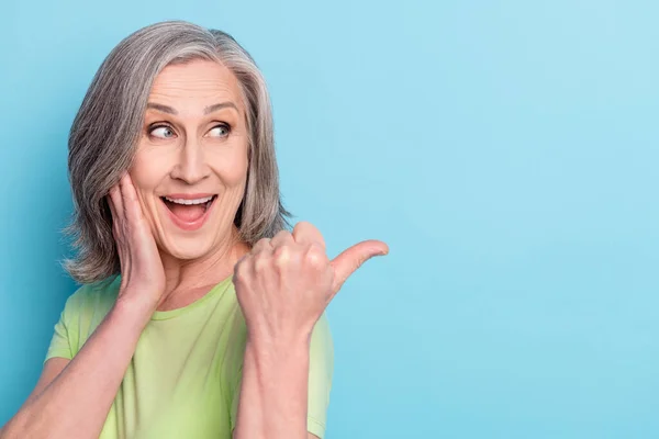 Foto van verbaasd positieve oude vrouw wijzen vinger lege ruimte houd hand wang geïsoleerd op blauwe kleur achtergrond — Stockfoto