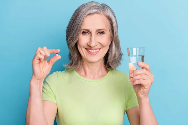 Foto de alegre velha senhora aposentada segurar as mãos pílula de vidro de água sorriso isolado no fundo de cor azul — Fotografia de Stock