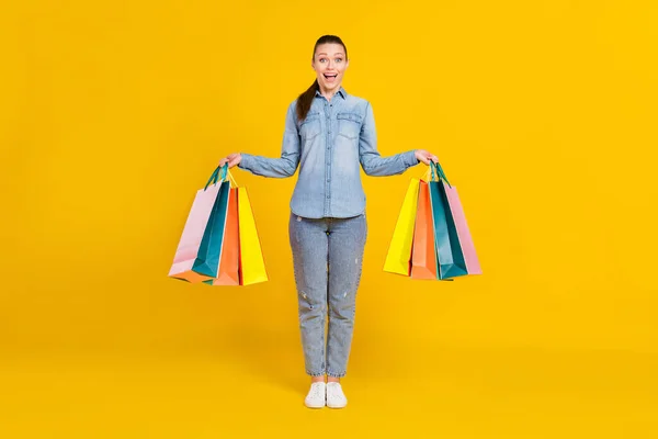 Full length photo of adorable impressed woman wear denim shirt smiling holding shopping bags isolated yellow color background — Stock Photo, Image