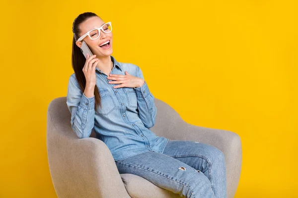 Zdjęcie słodkie śmieszne kobieta nosić dżinsowe okulary koszula śmiech siedzi krzesło rozmowy nowoczesny gadżet izolowane żółty kolor tło — Zdjęcie stockowe