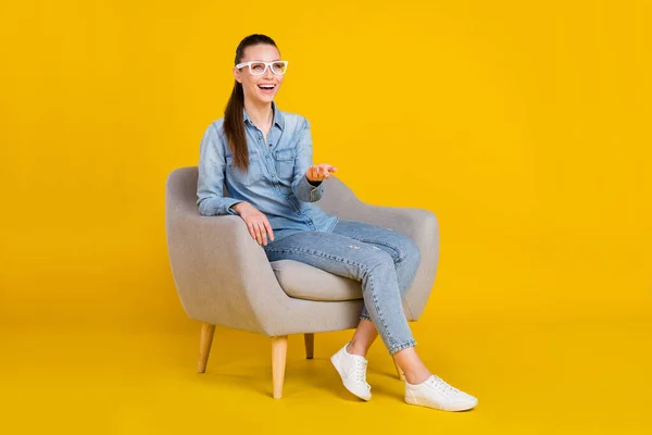 Photo of pretty shiny young lady dressed jeans shirt spectacles sitting chair looking empty space smiling isolated yellow color background — 图库照片