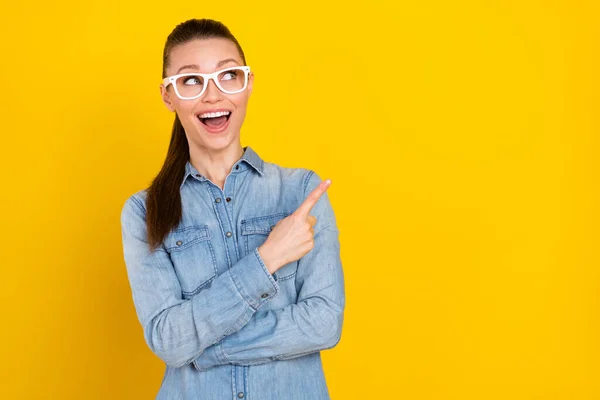 Foto de bonito impressionado jovem senhora vestida jeans camisa óculos apontando dedo espaço vazio sorrindo isolado cor amarela fundo — Fotografia de Stock