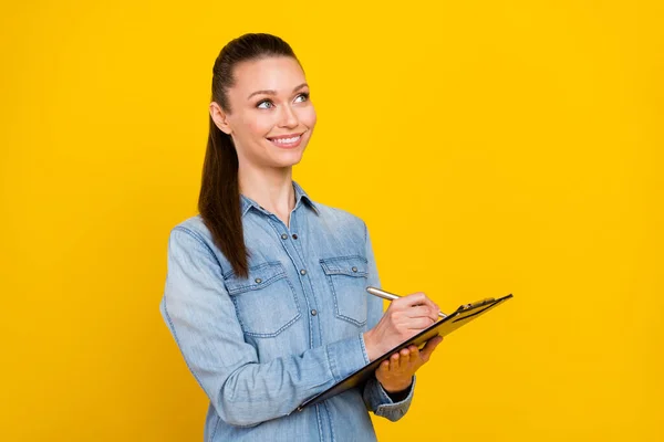 Foto de bonito doce jovem mulher usar camisa jeans sorrindo olhando espaço vazio escrita prancheta isolado cor amarela fundo — Fotografia de Stock