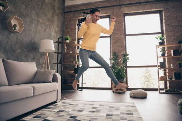 Foto de divertida dulce joven dama usar suéter amarillo levantamiento puño celebración de gadget moderno saltar en el interior de la habitación casa — Foto de Stock