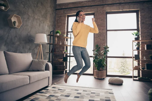 Foto de la joven afortunada brillante vestida suéter amarillo saltando hablando dispositivo moderno levantando puño en el interior habitación de casa —  Fotos de Stock