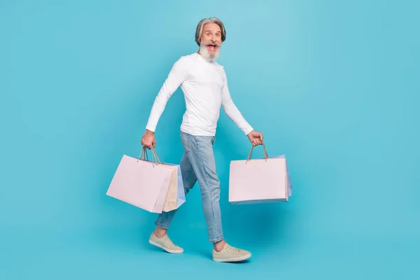 Volledige lichaamsomvang uitzicht van aantrekkelijke vrolijke grijs-harige man holding tassen gaan geïsoleerd over helder blauwe kleur achtergrond — Stockfoto