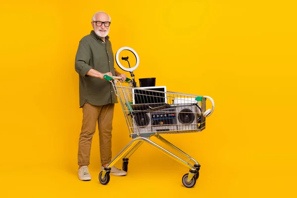 Full size photo of cheerful aged man push market trolley trade old stuff items isolated on yellow color background — 图库照片