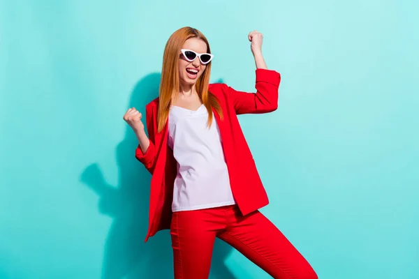 Photo of shiny lucky orange hair woman dressed red jacket dark eyewear rising fists smiling isolated cyan color background — Foto Stock