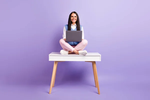 Retrato de la hermosa chica alegre morena sentada en la mesa en posición de loto utilizando el ordenador portátil que estudia aislado sobre el color violeta púrpura fondo — Foto de Stock