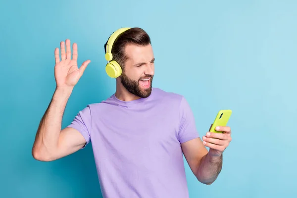 Fotoporträt eines bärtigen Mannes, der mit einem Handy im Kopfhörer auf hellblauem Hintergrund winkt — Stockfoto