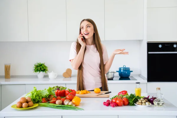 Портрет привабливої веселої мрійливої дівчини, яка готує смачну свіжу страву салат, слухаючи розмову на світло-білій домашній кухні в приміщенні — стокове фото