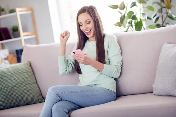 Foto wanita berambut cokelat hooray millennial duduk di sofa dengan telepon memakai celana jeans biru di rumah saja — Stok Foto