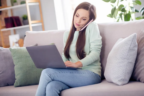 Фото милой блестящей молодой женщины, одетой в бирюзовый свитер, сидящей на диване и разговаривающей с современными устройствами, улыбающейся в помещении — стоковое фото