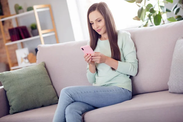 Photo of cute young brunette lady sit on sofa with telephone wear blue shirt jeans at home alone — Foto Stock