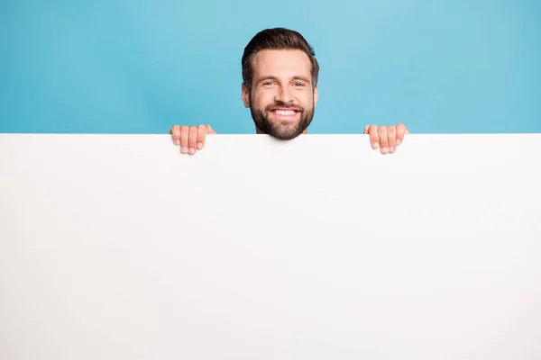 Foto retrato de divertido hombre juguetón escondido detrás de la pared blanca espacio en blanco para la promoción aislado sobre fondo de color azul brillante —  Fotos de Stock