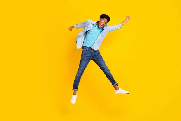 Photo of attractive lucky dark skin guy dressed denim shirt rising fists jumping high isolated yellow color background — Fotografia de Stock