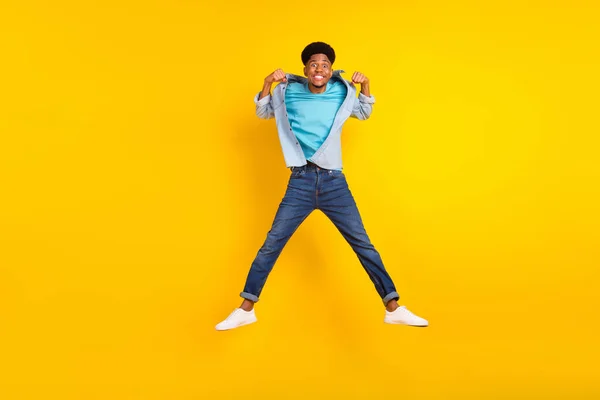 Foto de fuerte atractivo chico de piel oscura vestido camisa de mezclilla que muestra los músculos saltando alto aislado de color amarillo de fondo — Foto de Stock