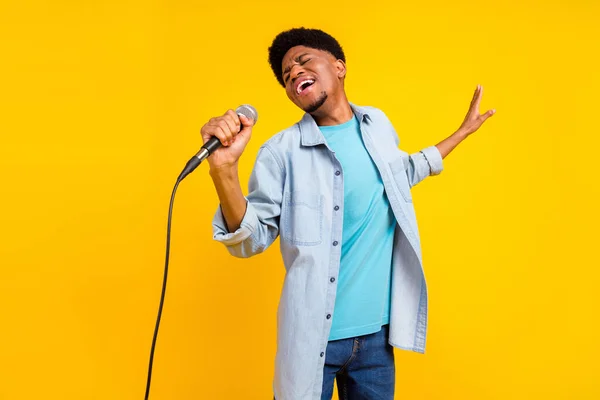 Foto de lindo atractivo chico de piel oscura vestido camisa de mezclilla cantando micrófono bailando aislado color amarillo fondo —  Fotos de Stock