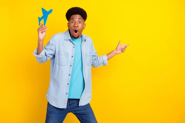 Foto de divertido impactado hombre de piel oscura usar jeans camisa sosteniendo avión de papel apuntando espacio vacío aislado color amarillo fondo — Foto de Stock