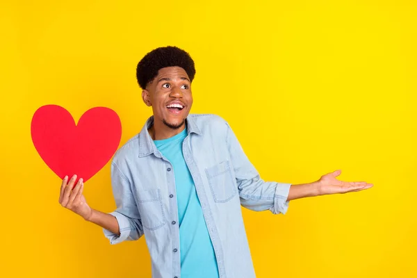 Foto de chico de piel oscura soñador impresionado vestido camisa de mezclilla buscando espacio vacío sosteniendo el corazón aislado color amarillo fondo —  Fotos de Stock