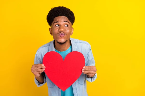 Foto de excitado lindo hombre de piel oscura usar jeans camisa sosteniendo grandes labios rojos del corazón pucheado buscando espacio vacío aislado color amarillo fondo — Foto de Stock