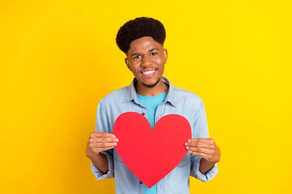 Foto de lindo encantador chico de piel oscura vestido camisa de mezclilla sonriendo sosteniendo gran corazón rojo aislado color amarillo fondo —  Fotos de Stock