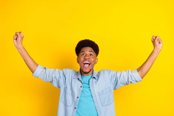 Photo of lucky pretty dark skin man wear jeans shirt holding rising fists looking up empty space smiling isolated yellow color background — Stock Photo, Image