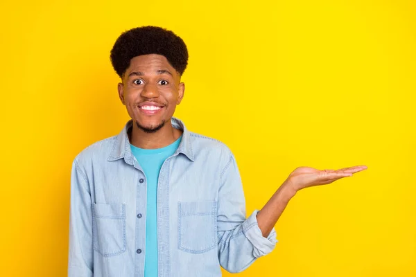 Photo of impressed pretty dark skin man wear jeans shirt holding arm empty space smiling isolated yellow color background — Stockfoto