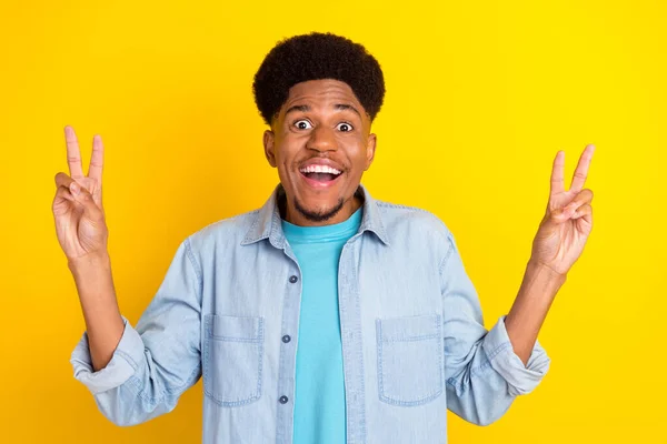 Foto van onder de indruk funky donkere huid guy gekleed denim shirt glimlachen met twee v-tekens geïsoleerde gele kleur achtergrond — Stockfoto