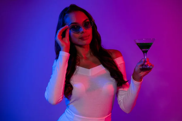Foto de dulce milenial morena señora beber alcohol con gafas de vestir blanco aislado sobre fondo de colores de neón —  Fotos de Stock