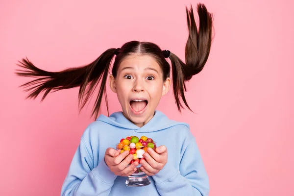 Foto retrato de colegiala impactada manteniendo el vidrio con dulces caramelos mirando con el pelo volador aislado sobre fondo de color rosa pastel —  Fotos de Stock