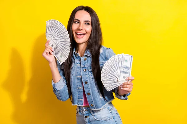 Фото милая милая женщина одета джинсовый наряд улыбаясь держа деньги вентиляторы выглядят пустое пространство изолированный желтый цвет фона — стоковое фото