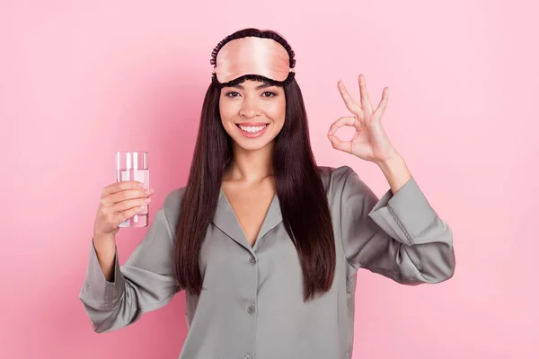 Фото очаровательной сияющей леди маска спального костюма питьевой воды, показывающей okey знак улыбаясь изолированный розовый цвет фона — стоковое фото