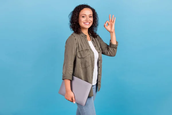 Retrato de menina de cabelos ondulados alegre atraente carregando laptop mostrando ok-sinal isolado sobre fundo de cor azul brilhante — Fotografia de Stock