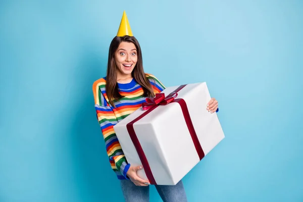 Retrato fotográfico de una chica sorprendida sosteniendo una gran caja de regalo envuelta con una gorra aislada sobre un fondo de color azul vivo —  Fotos de Stock