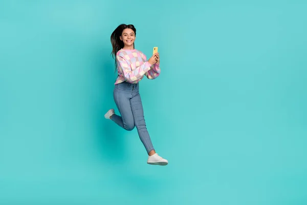 Parlak turkuaz rengi arkaplanda izole edilmiş bir cihaz uygulaması kullanarak atlayan güzel, neşeli kızın tam boy vücut büyüklüğü görünümü — Stok fotoğraf